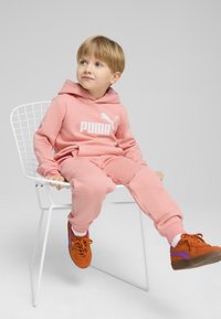 Rozā džemperis un bikses ar baltu PUMA logotipu, apvienotas ar oranžām kedām ar purpura akcentiem. Sēž uz baltas metāla krēsla.