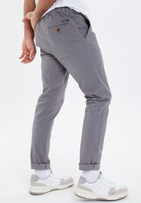 Light gray fitted trousers with an elastic waistband, rolled cuffs, and a back pocket featuring a button closure, paired with white sneakers.