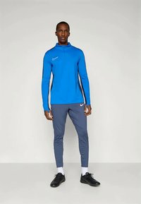 Blue long-sleeve athletic top with a high collar and zipper, paired with gray fitted pants. Both feature a white Nike logo. Black sports shoes.