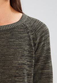 Stickad tröja i mörk olivgrön med ett strukturerat mönster, med en bred halsringning och raglanärmar, som visar upp varierade materialtexturer.