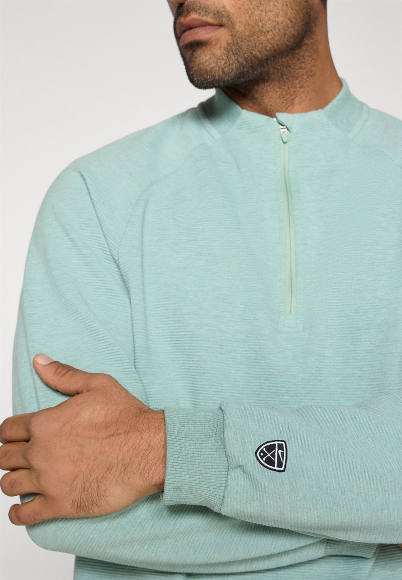 Pull-over bleu clair texturé avec un col à demi-fermeture éclair, des poignets côtelés et un petit logo noir brodé sur la manche.