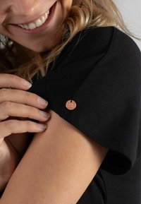 Schwarzes kurzärmliges Kleidungsstück mit glatter Textur und einem runden Kupfer-Logoanhänger, der am Ärmel befestigt ist. Nahaufnahme einer Hand auf dem Stoff.