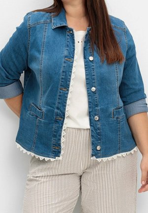 Frau trägt eine blaue Jeansjacke mit weißer Spitzenverzierung, ein weißes Oberteil und beige-weiß gestreifte Hosen, steht vor einem einfarbigen Hintergrund.