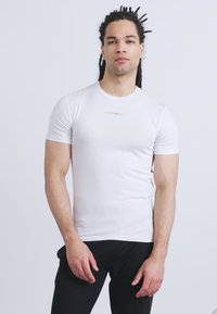 Άνδρας με dreadlocks, φορώντας εφαρμοστή λευκή μπλούζα και μαύρο παντελόνι, στέκεται μπροστά σε απλό ανοιχτό γκρι φόντο.