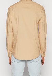 Camicia beige a maniche lunghe con taglio dritto, polsini abbottonati e una texture liscia. Presenta un dettaglio distintivo della cucitura dello yoke posteriore.