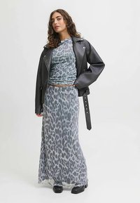 Žena s kudrnatými vlasy, oblečená v šedém topu s leopardím potiskem a odpovídající dlouhé sukni, černá kožená bunda přehozená přes ramena, stojící s rukou na bocích.