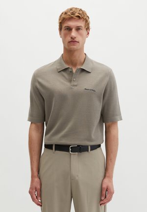 Mężczyzna w beżowej koszulce polo z logo Marc O'Polo, włożonej do beżowych spodni z czarnym paskiem, stojący na jednolitym tle.