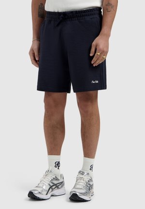 Male model wearing navy drawstring shorts, white socks with lettering, and silver running shoes, standing on a plain light gray background.
