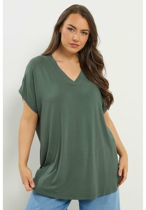 Frau mit langen braunen Haaren, die ein locker sitzendes dunkelgrünes V-Ausschnitt-Shirt mit kurzen Ärmeln und hellblaue Jeans vor einem schlichten weißen Hintergrund trägt.