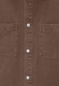 Bruine stof met twee vierkante zakken aan de voorkant en een centrale knopenlijst met vier ronde zilveren knopen.