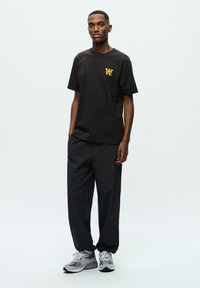 Black crew-neck t-shirt with yellow logo, paired with loose dark gray pants and gray athletic shoes with white accents. Simple design, casual fit.