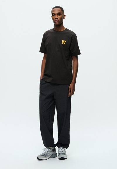Zwart crew-neck t-shirt met geel logo, gecombineerd met losse donkergrijze broek en grijze sportschoenen met witte accenten. Eenvoudig ontwerp, casual pasvorm.