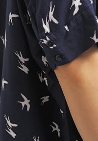 Chemise bleu marine avec motifs d'oiseaux blancs, manches courtes avec deux boutons noirs, tissu doux avec un léger éclat.