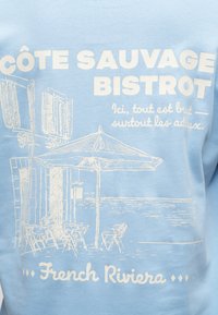 Μπλε ύφασμα με λευκό σχέδιο παραθαλάσσιου καφέ, ομπρέλα, τραπέζια, καρέκλες και κείμενο «Côte Sauvage Bistrot» και «French Riviera».