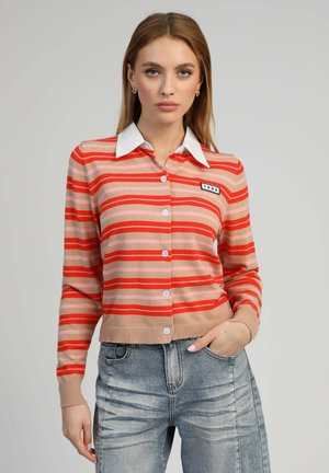 Jeune femme portant un cardigan à rayures rouge et beige boutonné avec un col blanc et un jean bleu délavé, posant devant un fond clair.