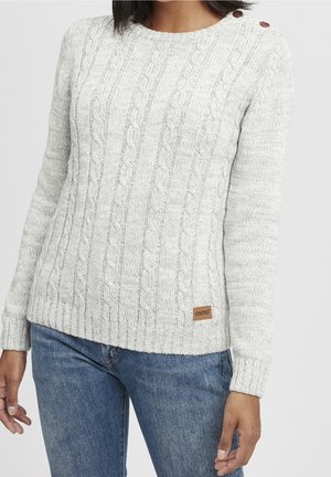 Femme portant un pull gris clair en maille torsadée avec des boutons aux épaules et un jean bleu, debout devant un fond uni.