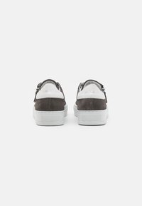 Sapatilhas de camurça castanha com detalhes brancos, de perfil baixo e sola de borracha texturizada. Apresentam um design slip-on com um fecho de correr na parte traseira.