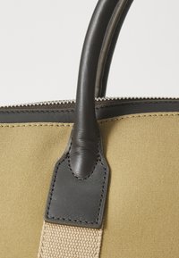 Olivenfarvet lærredstaske i nærbillede med mørkebrune læderhåndtag, udstyret med en lynlås og beige remaccenter, der fremhæver fine syninger.