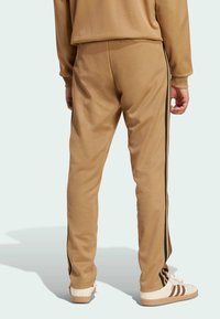 Light brown athletic pants with black side stripes and a relaxed fit, made from soft fabric. Worn with white sneakers featuring brown accents.