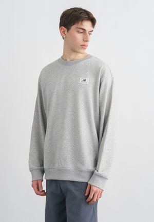 Nuori mies, joka on pukeutunut vaaleanharmaaseen collegepaitaan, jossa on pieni New Balancen logopläte, katsoo vasemmalleen yksiväristä taustaa vasten.