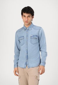 Camisa de denim azul claro con cuello puntiagudo, cierre de botones en el frente, dos bolsillos en el pecho y mangas largas con puños abotonados. Textura suave, ajuste clásico.
