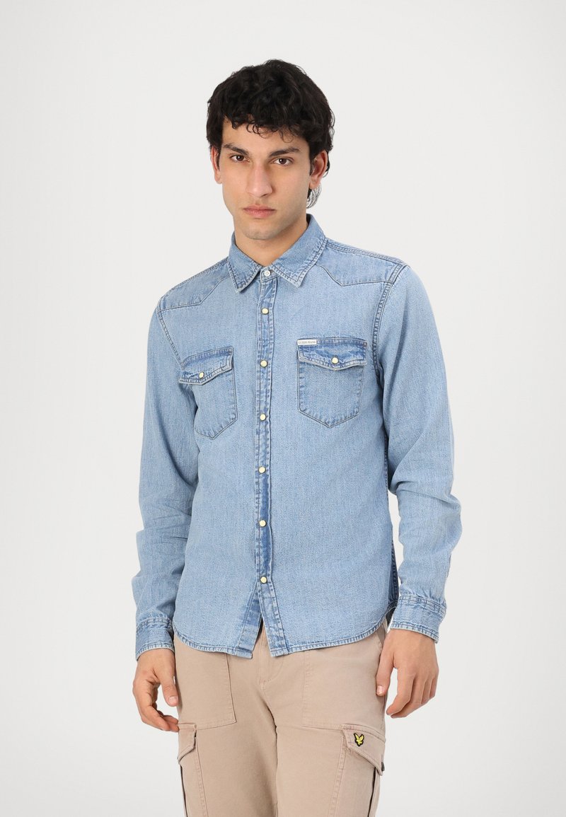 Camisa de denim azul claro con cuello puntiagudo, cierre de botones en el frente, dos bolsillos en el pecho y mangas largas con puños abotonados. Textura suave, ajuste clásico.