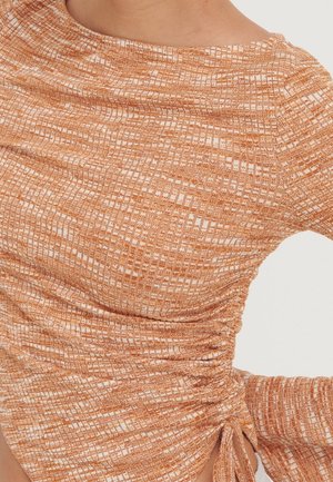 Gros plan d'une personne portant un haut texturé à manches longues avec un motif orange et blanc, avec un détail froncé sur le côté et un lien à la taille.