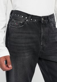 Schwarze Denim-Jeans mit geradem Schnitt, ausgestattet mit Metallnieten an der Taille, einer Vordertasche und einem kleinen Logo-Tag an der Tasche.