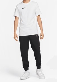 Bílé tričko Nike s černým logem, doplněné černými tepláky s logem a bílými teniskami s texturovaným designem.