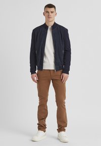 Marineblaues Wildleder-Bomberjackett, beige Pullover darunter, braune Hose, weiße Sneaker; glatte Textur, gerippte Bündchen und Reißverschlusstaschen.