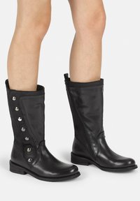 Botas de cuero negro hasta la rodilla con un cierre lateral, panel de tela y botones metálicos decorativos a lo largo del eje exterior. Suela de goma plana.