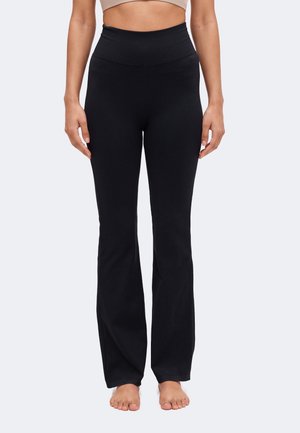 Un mannequin porte un legging noir évasé à taille haute et un crop top beige. Il se tient pieds nus sur un fond clair.