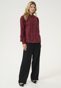 Blusa larga de manga larga en color burdeos con botones en el frente, diseño en capas y cuello tipo camisa, combinada con pantalones de pierna ancha negros y zapatos de tacón negro de punta.