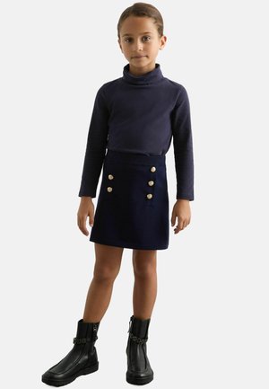 Marinblå polotröja i kombination med en mörkblå minikjol med guldknappar. Modellen bär svarta ankelboots med kedjedetalj.