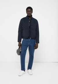 Veste matelassée navy avec un col haut, fermeture éclair à l'avant et deux poches inférieures ; associée à un jean bleu et des baskets blanches. Tenue d'un sac gris.