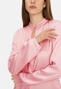 Rosa Satinbluse mit hohem Kragen, Knopfdetails und leichter Plissierung. Lange Ärmel mit Manschetten und Knopfverschlüssen.