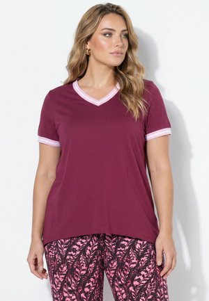 Donna che indossa una maglietta bordeaux con scollo a V e rifiniture bianche e pantaloni rosa con fantasia, in piedi davanti a uno sfondo bianco.