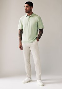 Polo a maniche corte verde chiaro con patta a tre bottoni, abbinata a pantaloni bianchi svasati e sneakers bianche. Tessuto morbido, vestibilità casual.