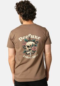 Man met krullend haar in een bruine T-shirt die het achterkantontwerp toont van een schedel met een dolk, rozen, een slang en de tekst "Deeluxe Tattoo Shop."