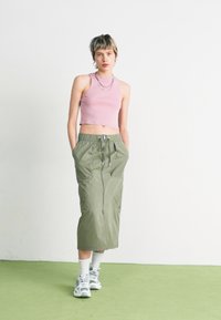 Világos rózsaszín bordázott crop top és egy olívazöld hosszú szoknya, cipzárral és zsebekkel, fehér zoknikkal és szürke sportcipőkkel párosítva.
