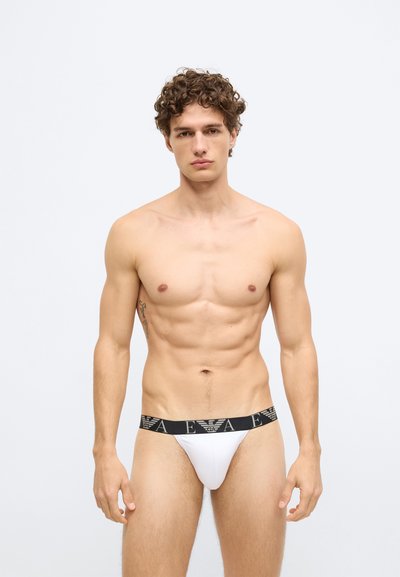 Biele slipy s čiernym elastickým pásom s detailom loga, priliehavý dizajn a hladká textúra látky. Model zobrazuje predný pohľad.