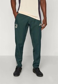 Zielone spodnie sportowe o lekko zwężanym kroju, z beżowymi logo adidas i Juventus oraz białymi paskami po bokach nóg.