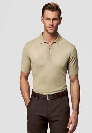 Mężczyzna ubrany w beżową, krótkorękawową koszulkę polo wpuszczoną w brązowe spodnie z ciemnobrązowym paskiem, stoi na tle jednolitego jasnego tła.