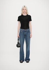 T-shirt nera aderente, jeans blu a taglio dritto e borsa nera con dettagli hardware. Modella in piedi su uno sfondo semplice.