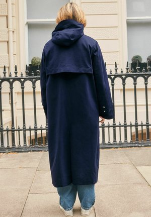 Persona con capelli biondi che indossa un lungo cappotto blu navy con cappuccio, jeans blu e scarpe bianche, in piedi su un marciapiede di città con la schiena rivolta.