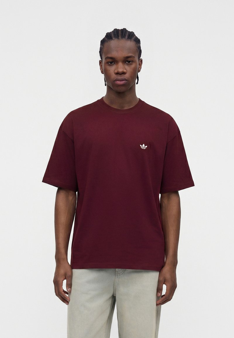 Junger Mann mit geflochtenem Haar, der ein schlichtes bordeauxrotes Adidas-T-Shirt und hellgraue Hosen trägt, steht vor einem weißen Hintergrund.