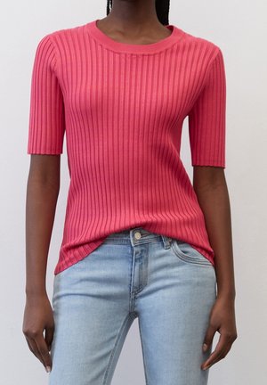 Vrouw die een nauwsluitende, geribde, roze top met korte mouwen draagt, licht ingestopt in lichtblauwe spijkerbroek tegen een effen achtergrond.