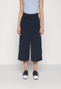 Navy culotte trousers with a tied waistband, made from a smooth fabric. Paired with black sneakers, featuring a low-top design and laces.