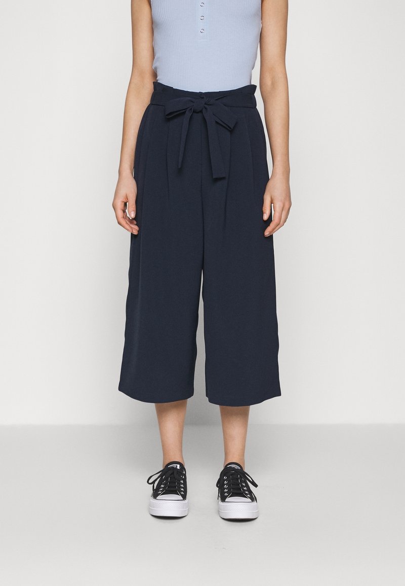 Navy culotte trousers with a tied waistband, made from a smooth fabric. Paired with black sneakers, featuring a low-top design and laces.