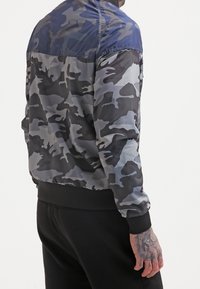 Mężczyzna w niebiesko-szarym kamuflażowym kurtce z czarnymi ściągaczami przy mankietach i w pasie, w połączeniu z czarnymi spodniami, widoczny tatuaż na dłoni.